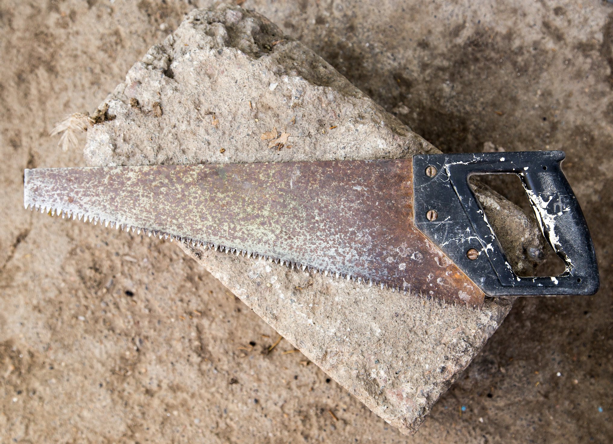 Close-up of an old hand saw with a rusted metal blade and worn black handle, resting diagonally on a rough concrete block
