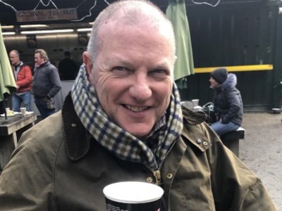 John Cushion wearing a checked scarf and raising a cup of coffee to the camera