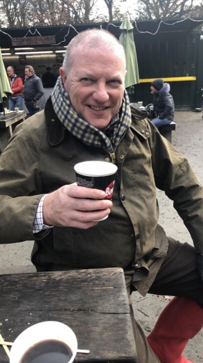 John Cushion wearing a checked scarf smiling and raising a coffee cup to the camera