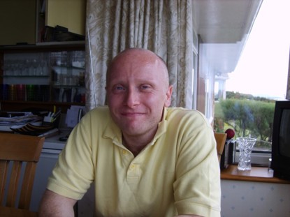 Dan Kay: a white bald man smiling at home wearing a yellow polo shirt