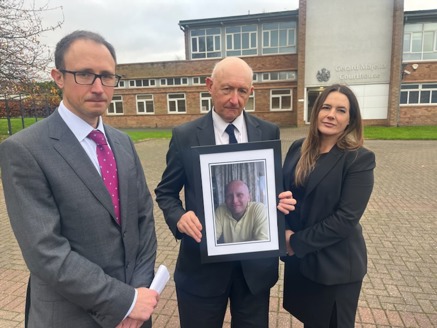 Three people wearing suits standing outside a building holding a framed picture of Dan Kay