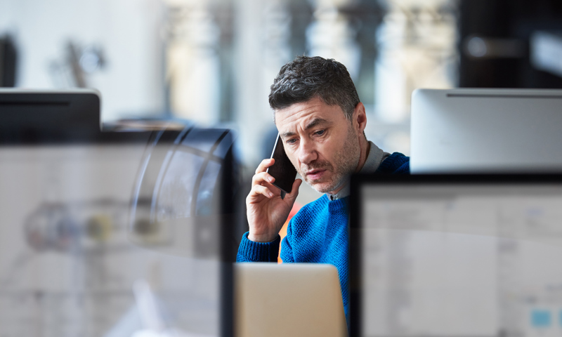 Man in blue sweater on phone, hidden amongst computer screens