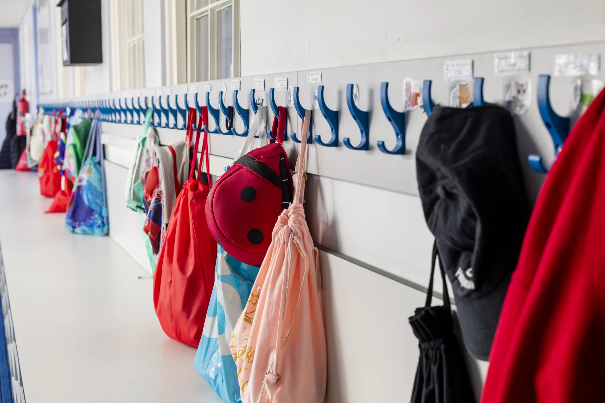 A school corridor lined with blue coat hooks holding colourful children's bags, backpacks and hats.