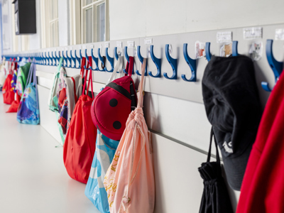 A row of blue clothes pegs on a white wall with red, blue and pink children's school bags hanging 