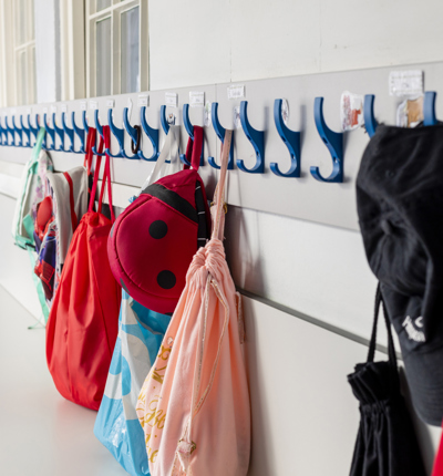A row of blue clothes pegs on a white wall with red, blue and pink children's school bags hanging 