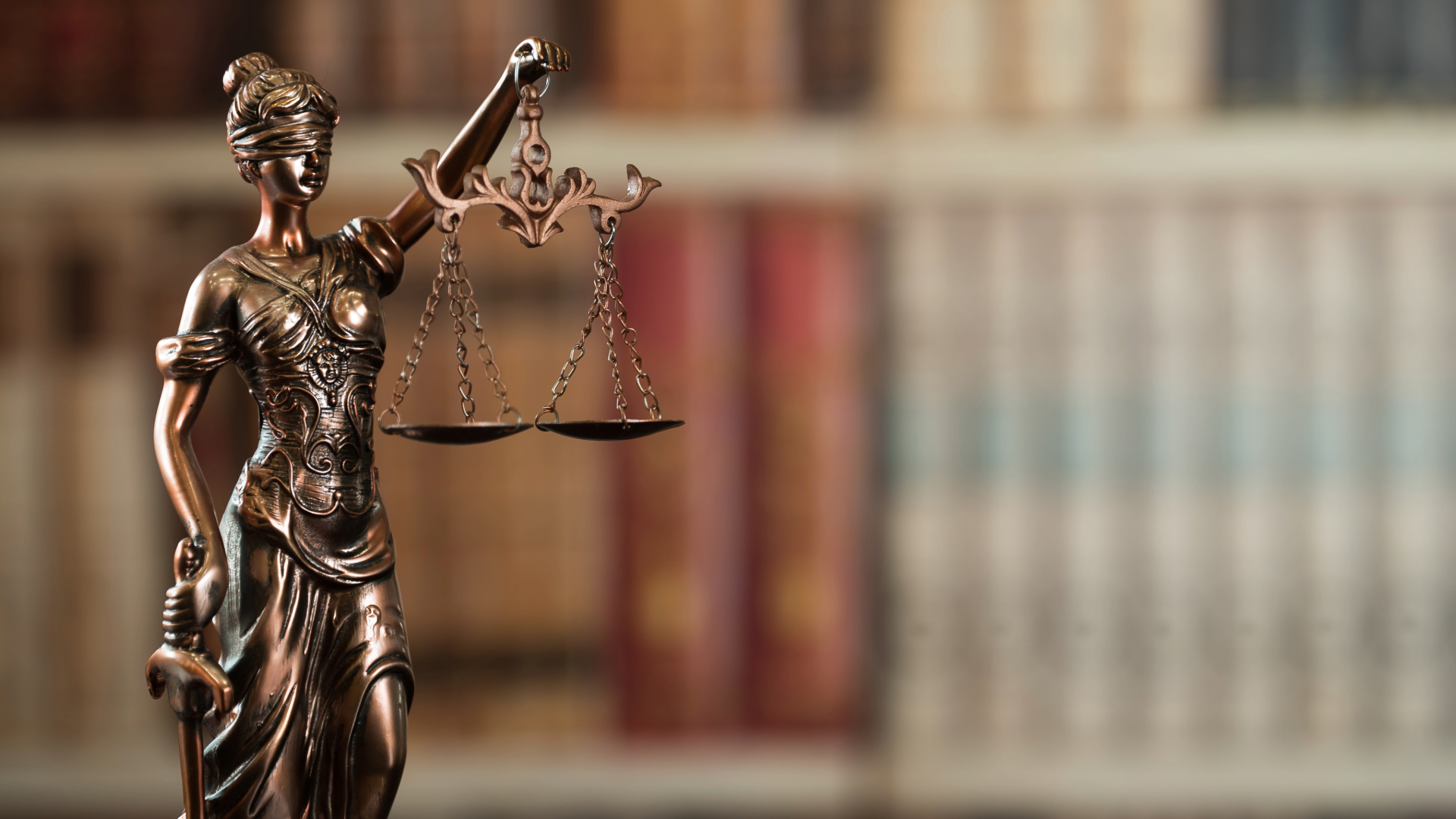Bronze Lady Justice statue holding balanced scales, with blurred bookshelves in the background