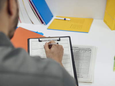 An over the shoulder photograph of a doctor in a grey shirt filling out a form on white paper on a black clipboard
