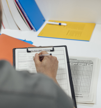 An over the shoulder photograph of a doctor in a grey shirt filling out a form on white paper on a black clipboard