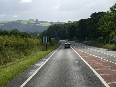 A grey concrete road with a blue car and green hedges and trees with grey blue sky