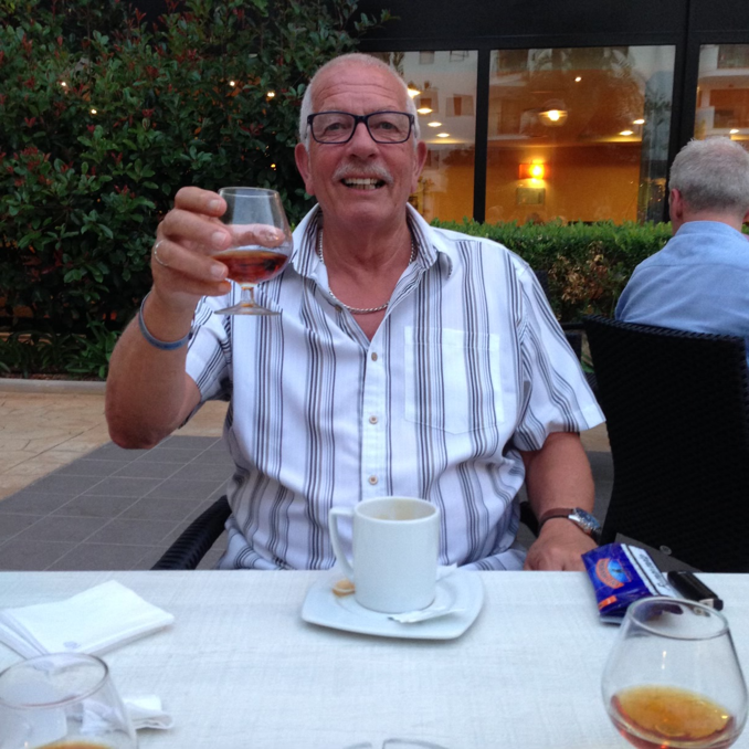 Geoffrey King sitting at a table smiling and raising a glass to the camera