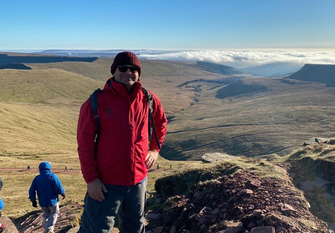 Man wearing red coat and hat smiling on top of a hill in Wye valley